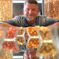 Michael Althoff sitzt hinter verschiedenen Popcornsorten in kleinen Schalen und blickt in die Kamera, während im Hintergrund große Popcornbehälter stehen.