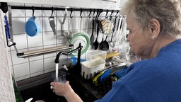 Eine Frau steht vor einer Spüle in einer Küche und lässt Wasser aus dem Wasserhahn in ein Glas fließen