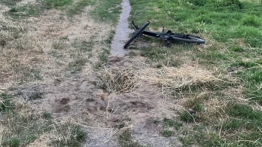 Auf einem Feldweg ist eine Mulde mit Stroh und Gras verdeckt, dahinter liegt ein Fahrrad auf dem Boden