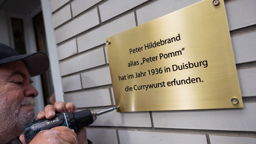 Ein Mann schraubt eine goldene Plakette an einer Hauswand an. 