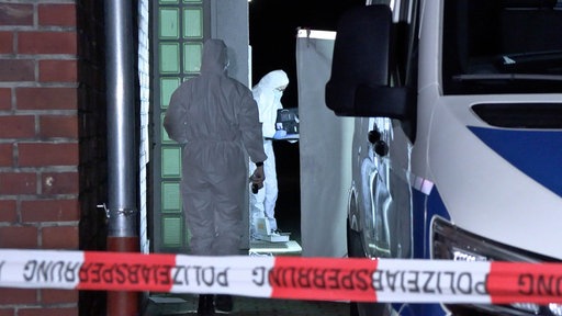 Zwei Personen in weißen Schutzanzügen neben einem Polizeiauto, davor ein Absperrband "Polizeiabsperrung"