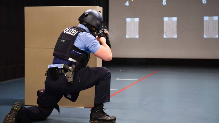 Eine Person mit Helm und Weste der Polizei, kniet vor zwei Karton und zielt mit einer Waffe auf Ziele