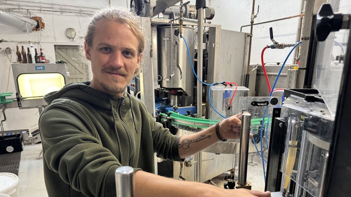 Christian Vormann, ein junger Mann mit langen Haaren und Bart, steht in der Brauerei und lächelt in die Kamera.