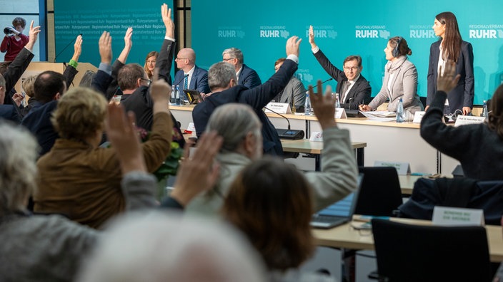 Menschen sitzen sich in einem Saal gegenüber, einige heben die Hände. In Hintergrund eine Leinwand auf der "Regionalverband Ruhr" steht.