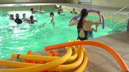 Am Poolrand liegen Poolnudeln, dahinter sieht man den Schwimmkurs beim Unterricht im Wasser. 