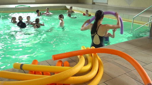 Am Poolrand liegen Poolnudeln, dahinter sieht man den Schwimmkurs beim Unterricht im Wasser. 