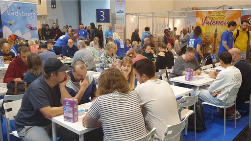 An mehreren Tischen in einer Halle sitzen Personen und spielen Spiele