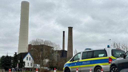 Ein Polizeiauto steht auf der Straße, Absperrungen durch Warnkegel auf der Straße. Im Hintergrund drei große Kraftwerksschornsteine.