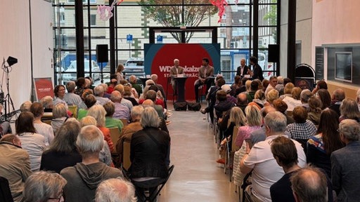 Viele Menschen sitzen in einer Halle in Stuhlreihen nebeneinander mit dem Rücken zur Kamera, vorne im Raum mehrere Personen auf dem Podium und ein Schild "WDR Lokalzeit Stadtgespräch"