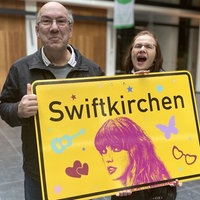 Ein Mann und eine Frau halten ein gelbes "Swiftkirchen"-Schild. Beide lachen und freuen sich. 