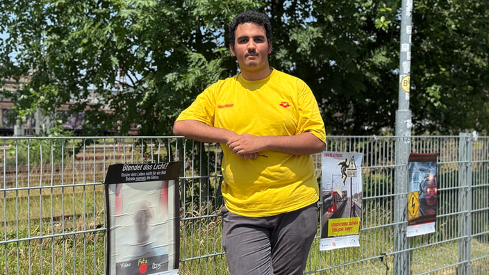 Nasser Mansour steht neben einem Zaun, an dem Plakate hängen. 