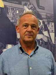 "2 Brüder"-Geschäftsführer Bart Rehe vor einem Plakat im Supermarkt, das die echten 2 Brüder zeigt 