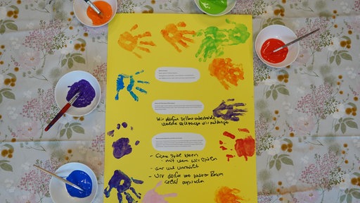 Ein gelbes Plakat mit Kinderrechten und bunten Handabdrücken liegt umringt von Schüsseln mit Farbe auf einem Bastelteppich.