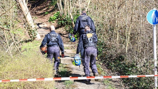 Drei Polizisten laufen hinter einem Absperrband einen Waldweg hinauf. 