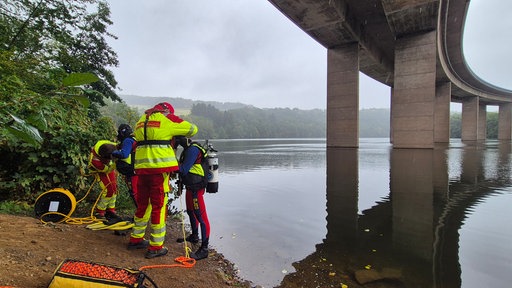 Einsatzkräfte stehen am Ufer unter einer Brücke