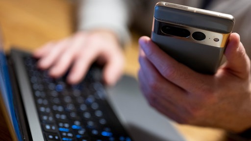 Eine person hält ein Handy in der Hand und tippt dabei auf einem Computer