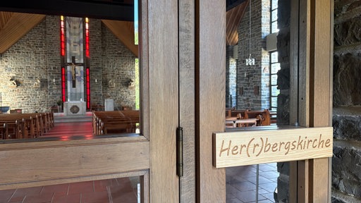 Ein Schild mit der Aufschrift "Her(r)bergskirche" hängt an einer Kirchentür