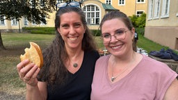 Zwei erwachsene Frauen stehen in einem Garten und lächeln in die Kamera. Die linke hat ein angebissenes Brötchen in der Hand.