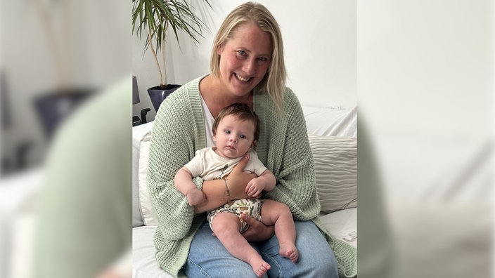 Eine blonde Frau sitzt auf einem Sofa, auf ihrem Schoß ein Kleinkind. Die Frau lacht in die Kamera.