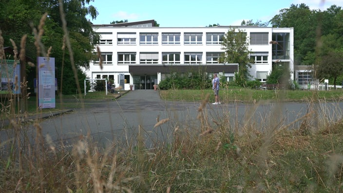 Ein weißes Gebäude, davor Büsche. Eine Person spaziert daran vorbei.
