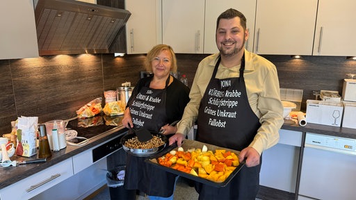 Eine Frau und eine Mann haben eine Schürze um, und präsentieren in der Küche eine Pfanne mit angebratenen Haselnüssen und ein Backblech mit Gemüse