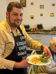 Ein Koch serviert einen Teller mit mit Haselnuss-Pesto und lächelt dabei