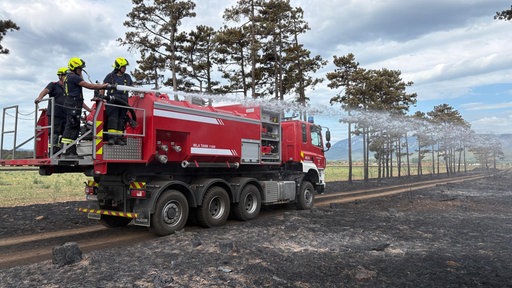 Feuerwehrleute spritzen von einem mobilen Wassertank aus Löschwasser auf verbrannt aussehende Fläche