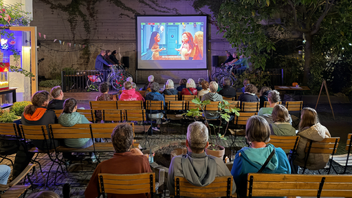 Auf einer Leinwand läuft ein Film. Rechts und links sitzen mehrere Menschen auf Fahrrädern.