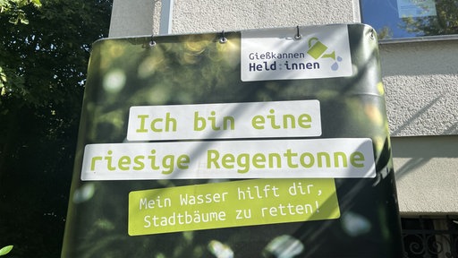 Regentonne mit der Aufschrift "Ich bin eine riesige Wassertonne. Mein Wasser hilft dir, Stadtbäume zu retten!"