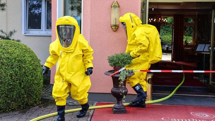 Zwei Menschen mit gelben Schutzanzügen vor einem abgesperrten Bereich