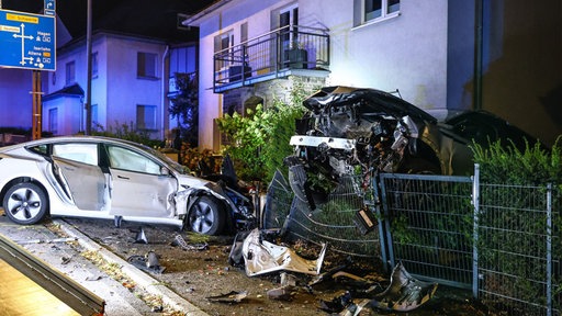 Zwei zerstörte Autos vor einem Haus, das eine hängt über einem Zaun