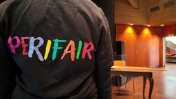 Eine junge Frau dreht der Kamera den Rücken zu: auf ihrem T-Shirt steht Perifair