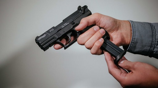 Eine männliche Hand umgreift eine schwarze Schreckschusswaffe, ein Finger ist neben dem Abzug, die andere Hand schiebt ein Magazin in die Waffe