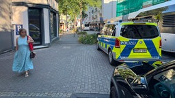 Polizeiauto steht in einer Einfahrt, Busse stehen in Schlange, Passantin schaut skeptisch auf das Geschehen