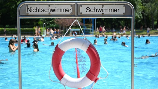 Vor einem Schwimmbecken steht ein Schild mit "Nichtschwimmer" und "Schwimmer", da drunter hängt ein Rettungsring, im Hintergrund sieht man Menschen im Wasser
