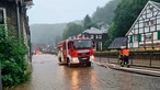 Auf einer überfluteten Straße in Solingen-Unterburg steht ein Feuerwehrwagen, von dem weiße Schläuche abgehen, an der Seite stehen zwei Feuerwehrmänner.