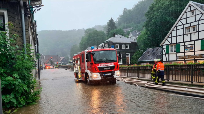 Auf einer überfluteten Straße in Solingen-Unterburg steht ein Feuerwehrwagen, von dem weiße Schläuche abgehen, an der Seite stehen zwei Feuerwehrmänner.