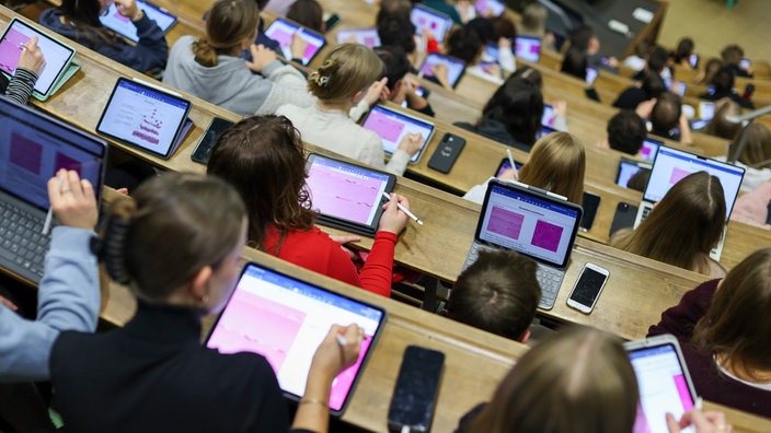 Studierende sitzen in einem Hörsaal einer Universität und schreiben auf ihren Tablets und Laptops