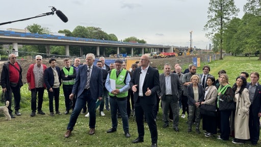 Eine Menschengruppe steht auf einer Wiese, im Hintergrund eine Brückenbaustelle. Im Vordergrund drei Männer, die gestikulieren. Ein Mikrofon ragt aus der linken oberen Bildecke rein.