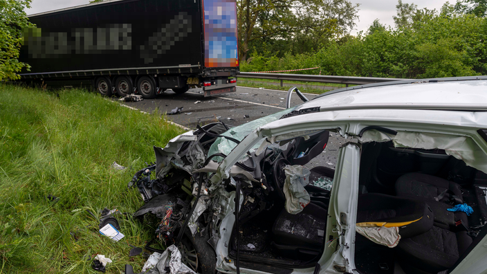 Ein stark beschädigtes weißes Fahrzeug steht an einem Straßenrand. Auf der Straße steht ein blauer LKW sowie herumliegende Fahrzeugteile.