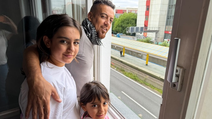 Ibrahim, Shavi und Lisa stehen in ihrer Wohnung am geöffneten Fenster, darunter verläuft die leere A40