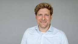 Ein Mann im Hemd lächelt vor neutralen Grund in die Kamera.