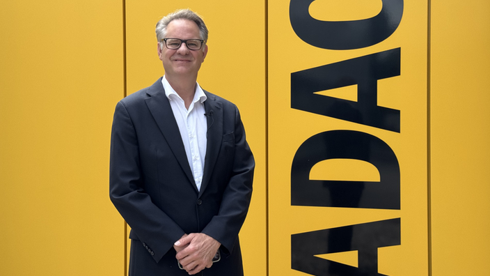 Roman Suthof steht im Anzug vor einer gelben Wand mit der Aufschrift ADAC