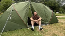 Ein Mann mit kurzer Hose und T-Shirt sitzt im Eingang eines grünen Zelts