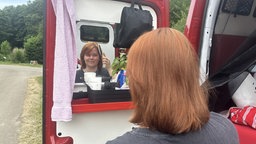 Eine Frau mit roten Haaren blickt in einen Spiegel in der Hecktüre eines Transporters und kämmt sich die Haare