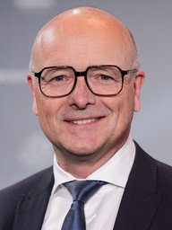 Karsten Schwanke lächelt freundlich in die Kamera. Er trägt einen Anzug mit Krawatte und eine Brille. 