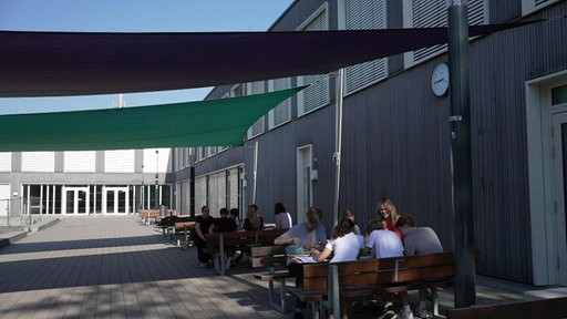 Auf einem Schulhof sitzen Schülern an Tischen unter Sonnensegeln
