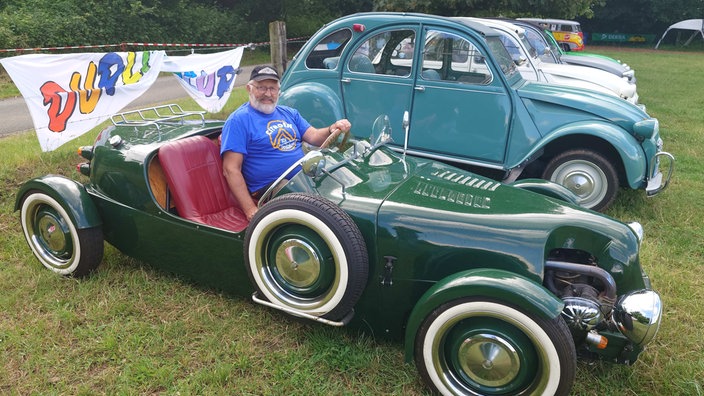 Ein älterer Mann mit Brille, weiß/grauem Bart und Haaren sitzt in einem dunkelgrünen Oldtimer, dahinter stehen weitere Oldtimer-Fahrzeuge