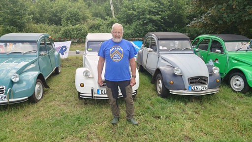 Ein älterer Mann mit weiß/grauem Bart und weißen Haaren steht in blauem Shirt vor vier alten Oldtimer-Fahrzeugen (Enten)