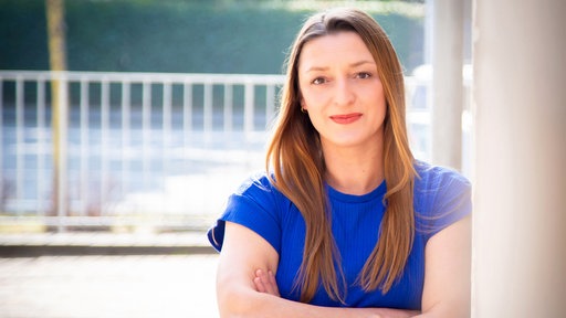 Edith Stecker lächelt in die Kamera. Sie hat braune Haare und ein blaues Oberteil und steht angelehnt an eine Wand.